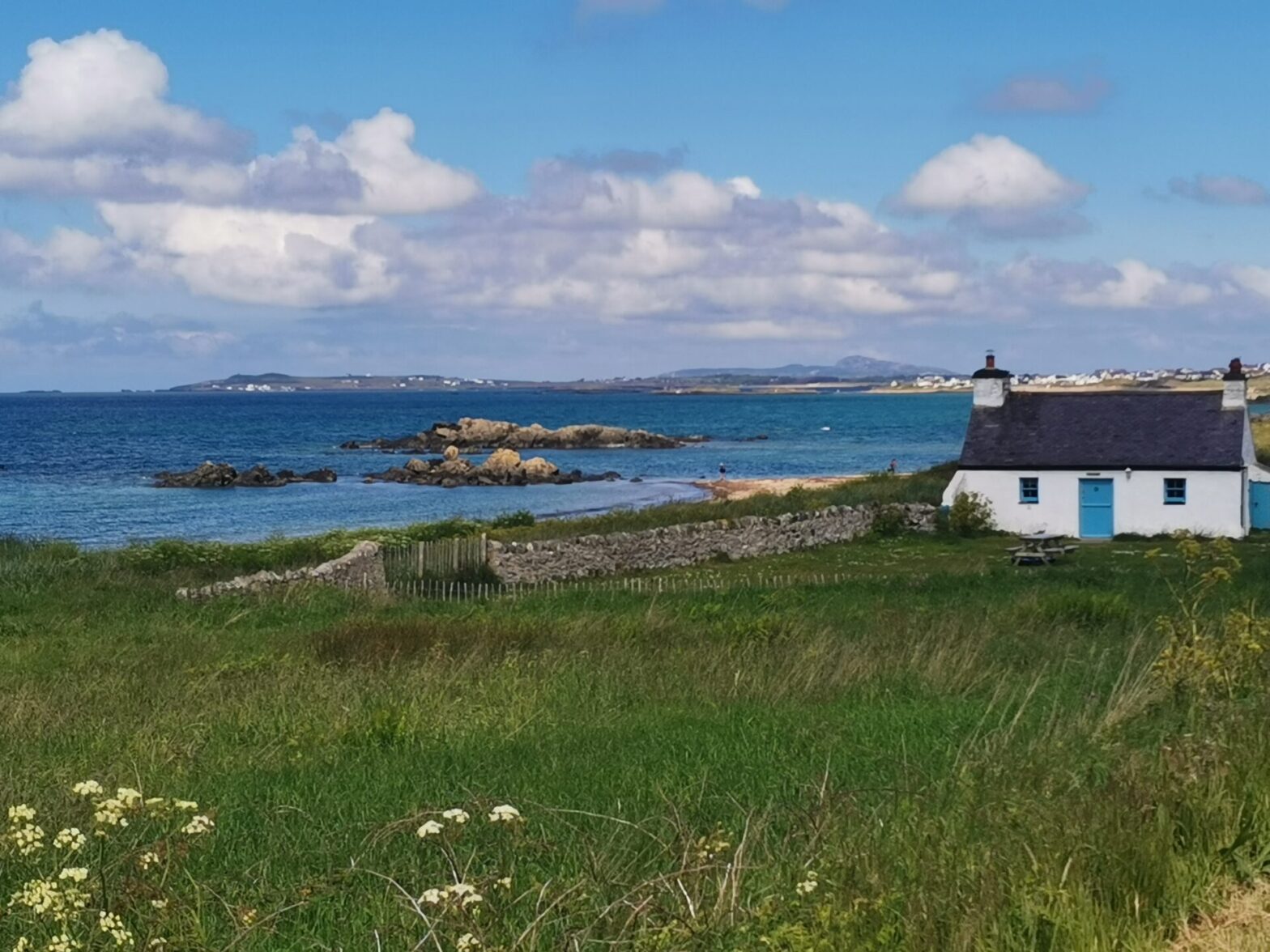 A pretty white cottage with a blue door stands at the endge of a rocky shore and blue sea on Anglesey