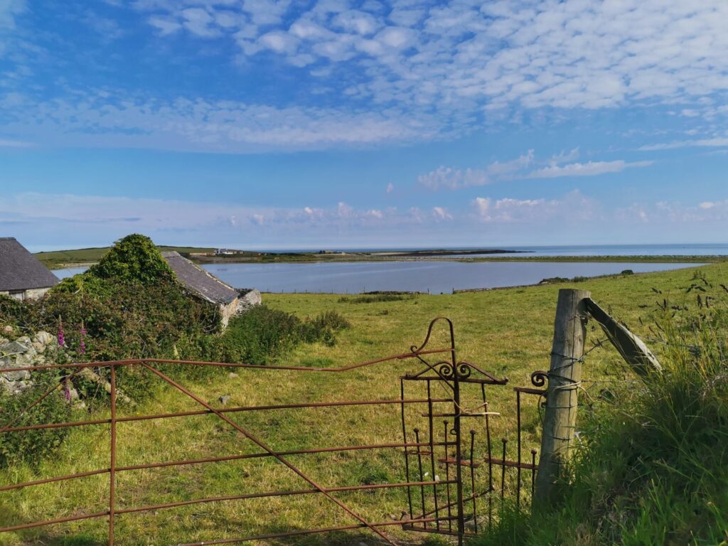 Looking over a rusty old farm gate and past farm buildings towards a lagoon, and sea, Anglesey