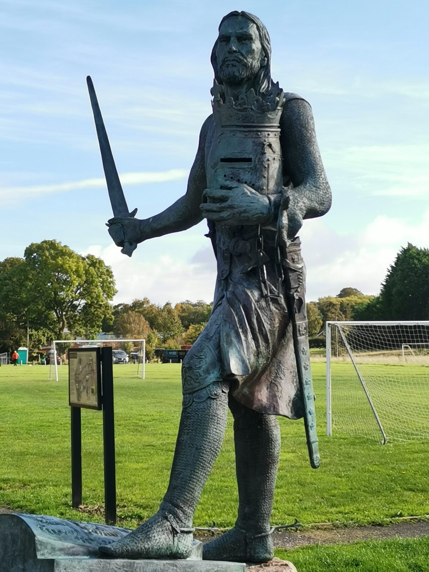 A statue of a medieval king in chain mail stands at the side of a village football ground.